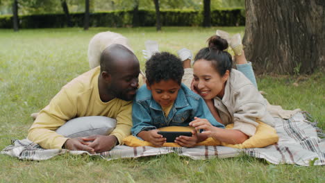 cheerful african american family lying in park and using smartphone