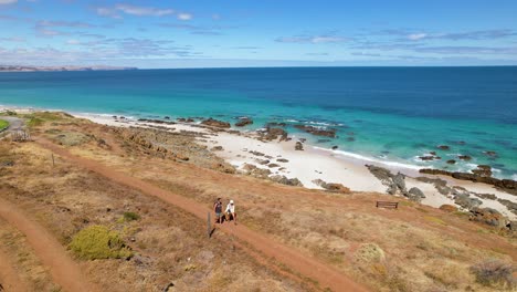 A-couple-walking-hand-in-hand-along-a-dirt-path-in-a-remote-landscape-with-a-bright-blue-ocean-and-white-sand-beach-next-to-them