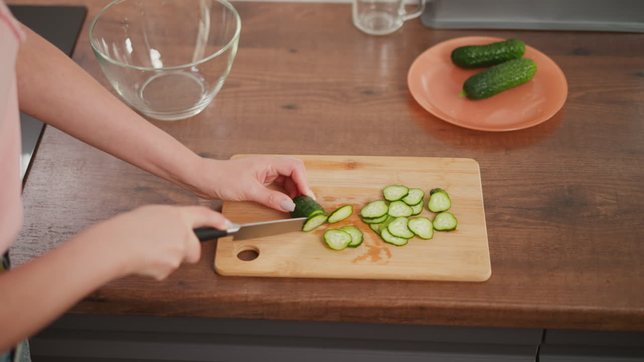 Close up of girl slicing cucumber into thin round pieces on wooden cutting board in kitchen, with two whole cucumbers on plate nearby and glass bowl resting on clean wooden counter