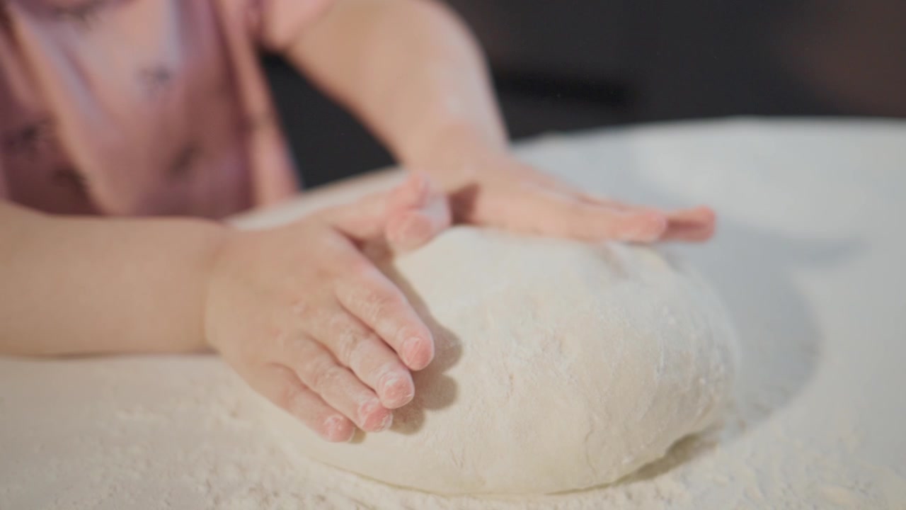 Close-up hands knead soft dough on floured surface. Child's hands press down gently, showing focus and care. Scene captures calm, creative moment in home kitchen. Ideal for cooking tutorials