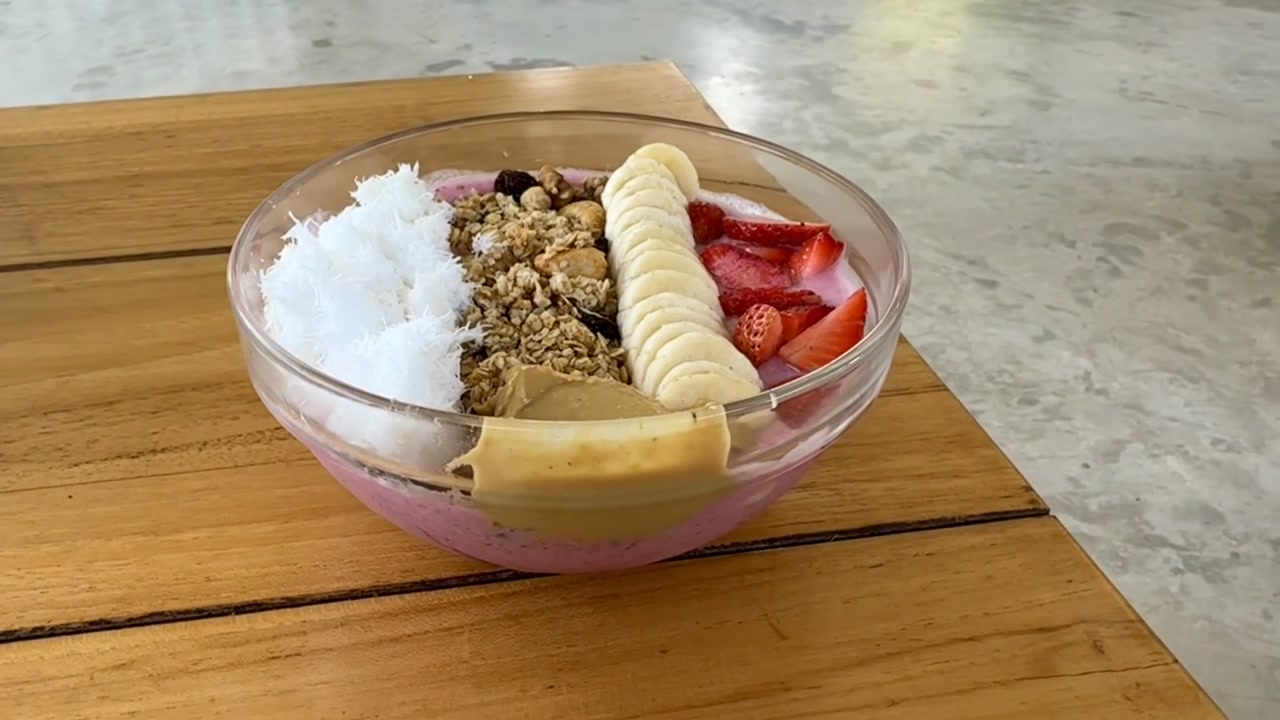 Riprese cinematografiche di cibo che mostrano una smoothie bowl vegana al frutto del drago, guarnita con banana, fragole, scaglie di cocco, granola fatta in casa e burro di arachidi cremoso.