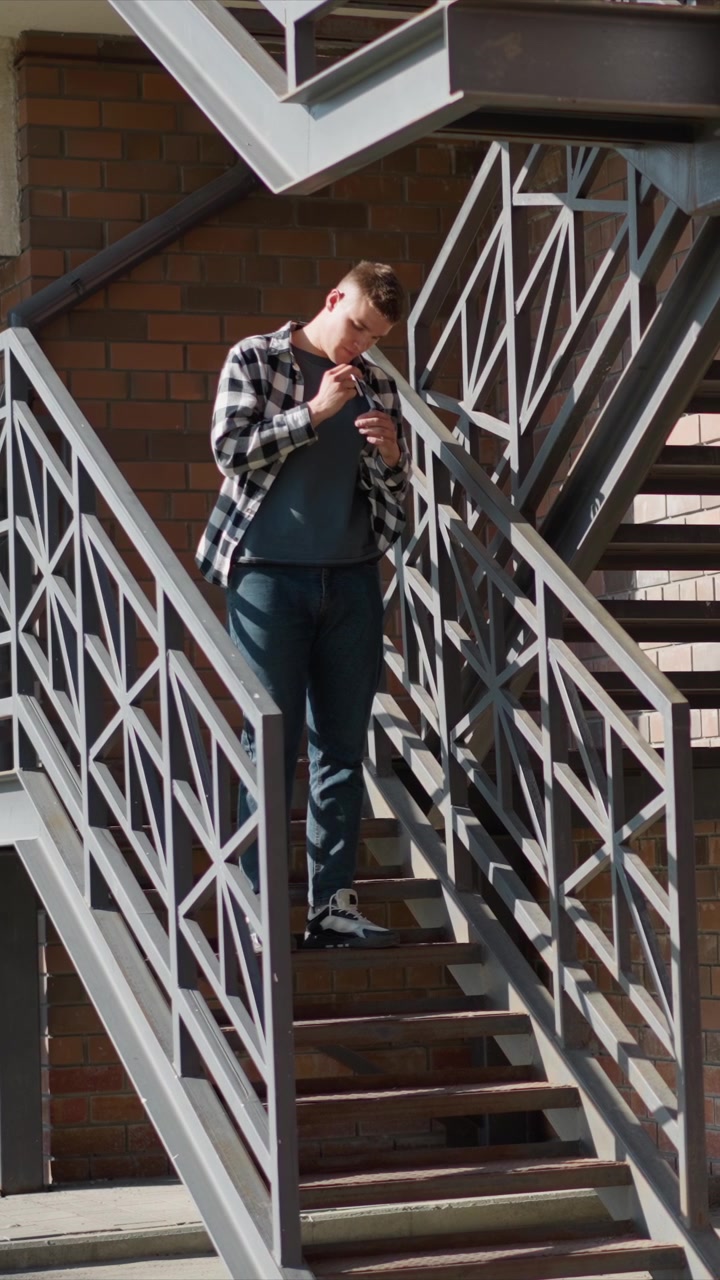 Nahaufnahme eines jungen Mannes, der im Freien eine Metalltreppe hinabsteigt, ein Mobiltelefon hält und benutzt. Der Mann trägt ein kariertes Hemd über einem T-Shirt mit Jeans und Turnschuhen vor einem Ziegelwandhintergrund. Sanfte Beleuchtung.