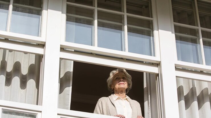 Sunlight drawing look, senior woman resting hands on open sash and watching outside at paned window