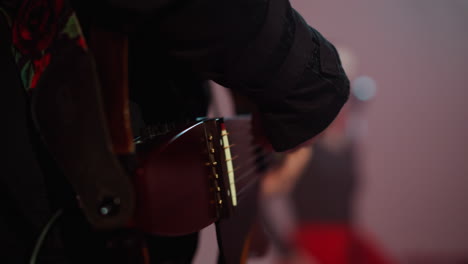 close-up of a guitarist's hand strumming a red guitar, with a blurred silhouette of a singer in the background, set against a softly focused colorful backdrop