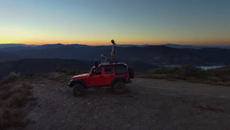 Aerial-circling-red-jeep-in-middle-of-nowhere,-man-climbs-up-roof-to-enjoy-view