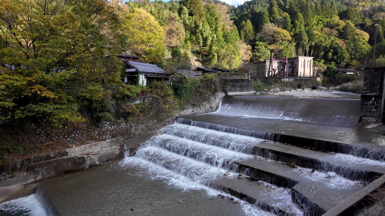 A tranquil river cascading over stepped weirs surrounded by lush forest and traditional buildings in Tsumago Post Town, showcasing peaceful nature and cultural charm.