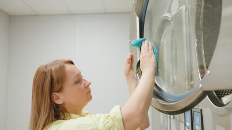 focused lady in yellow shirt wipes glass door of washing machine with soapy towel, carefully cleaning laundry equipment surface inside laundromat under bright light, ensuring cleanliness