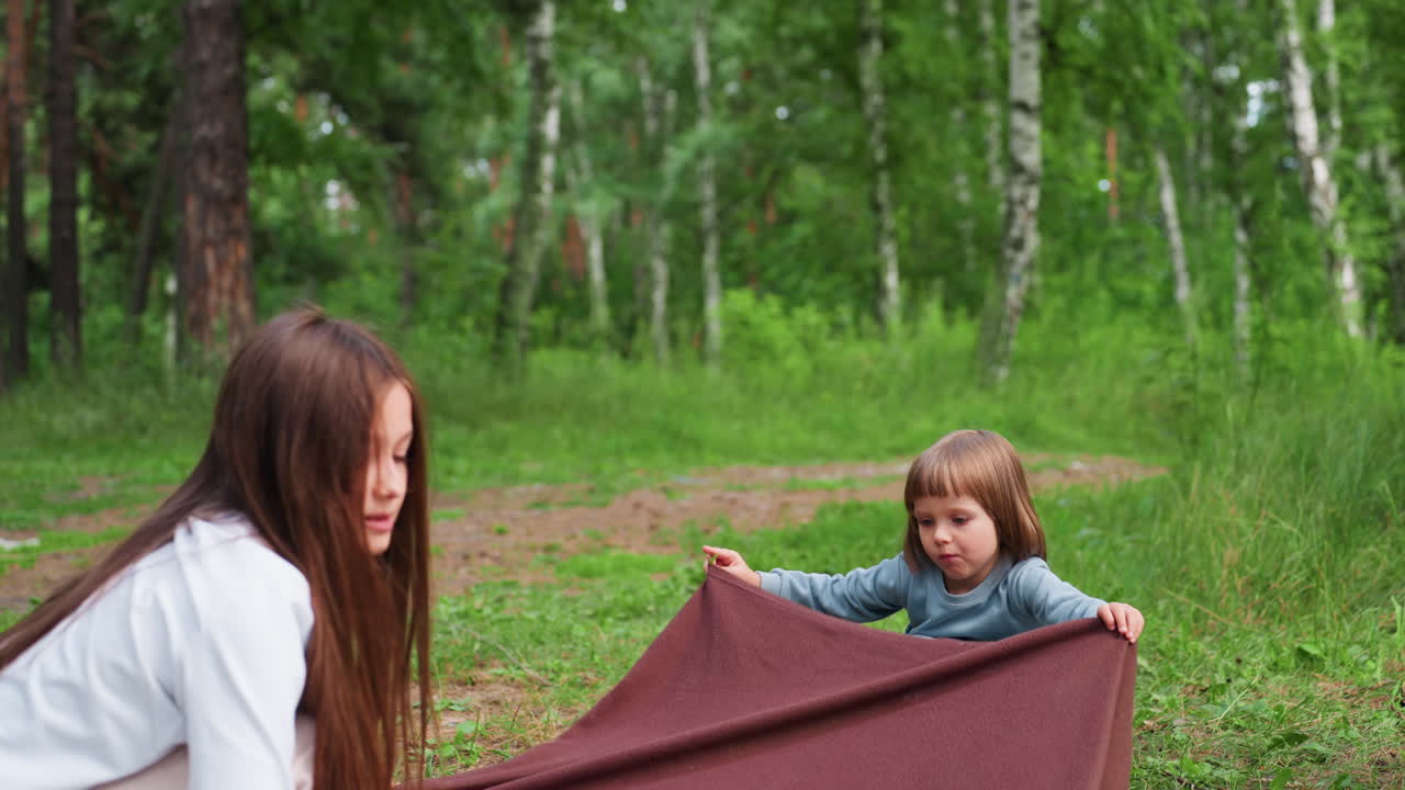 Две девочки расстилают коричневую скатерть для пикника на зеленой траве на тихой лесной поляне. Солнечный свет пробивается сквозь деревья, создавая радостный семейный момент, наполненный весельем, смехом и теплом на природе.