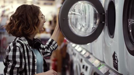 una mujer joven positiva en una camisa a cuadros eligiendo la lavadora en el tiro de los electrodomésticos. abre la puerta y mira