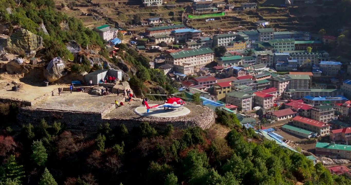 Namche Bazaar, Everest, Nepal. Yolcuların indiği tepe helikopter pistindeki inmiş bir helikoptere yukarıdan bakan drone çekimi, renkli yamaç evlerini ve yeşil tepeleri gözler önüne seriyor.
