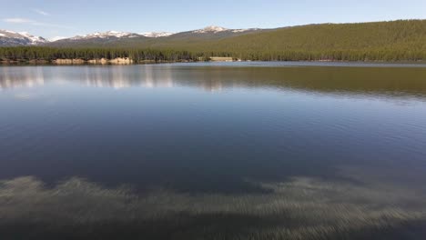 Forward-aerial-view-from-the-shore-to-the-lake-at-Park-Reservoir-in-summer-with-Clouds-Peak-Wilderness-in-the-background-in-Bighorn-National-Forest-in-Wyoming