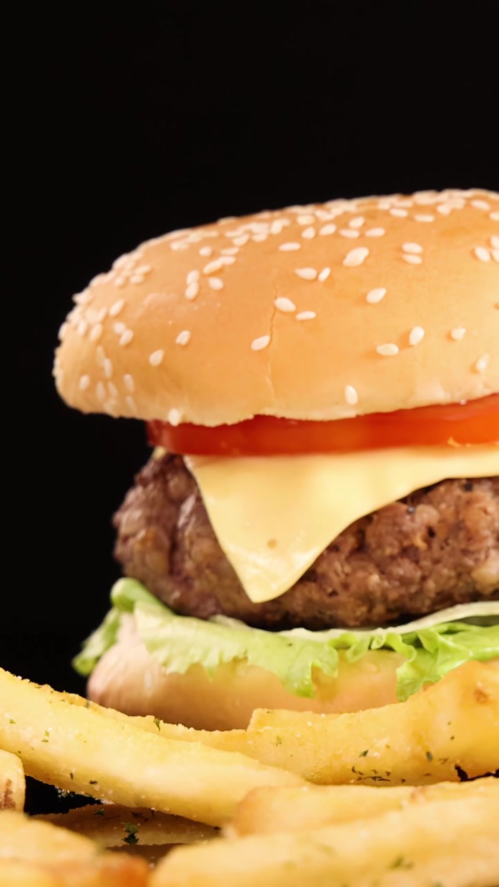 Patatas fritas doradas caen a cámara lenta junto a una hamburguesa con queso, lechuga, tomate y queso, bajo una brillante iluminación de estudio sobre un fondo negro