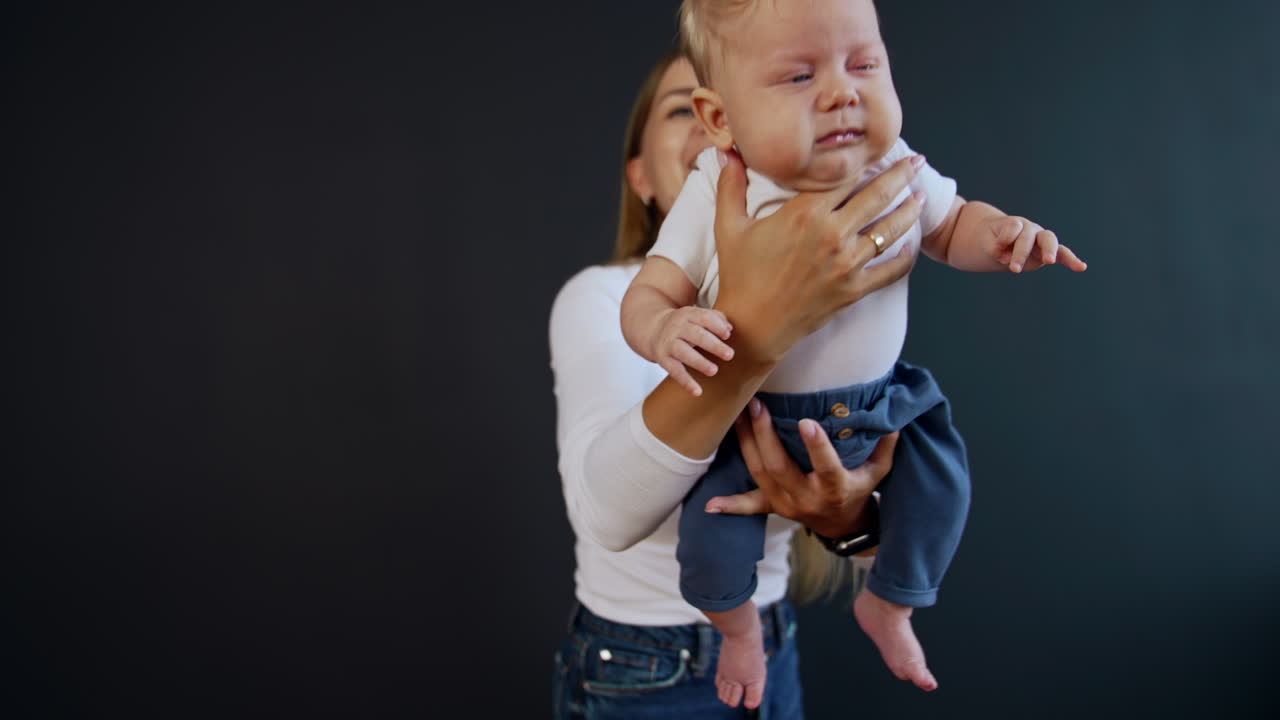 Blonde long-haired woman wearing white shirt and jeans holding a baby. Mother waves her baby and kid starts to cry. Black backdrop.