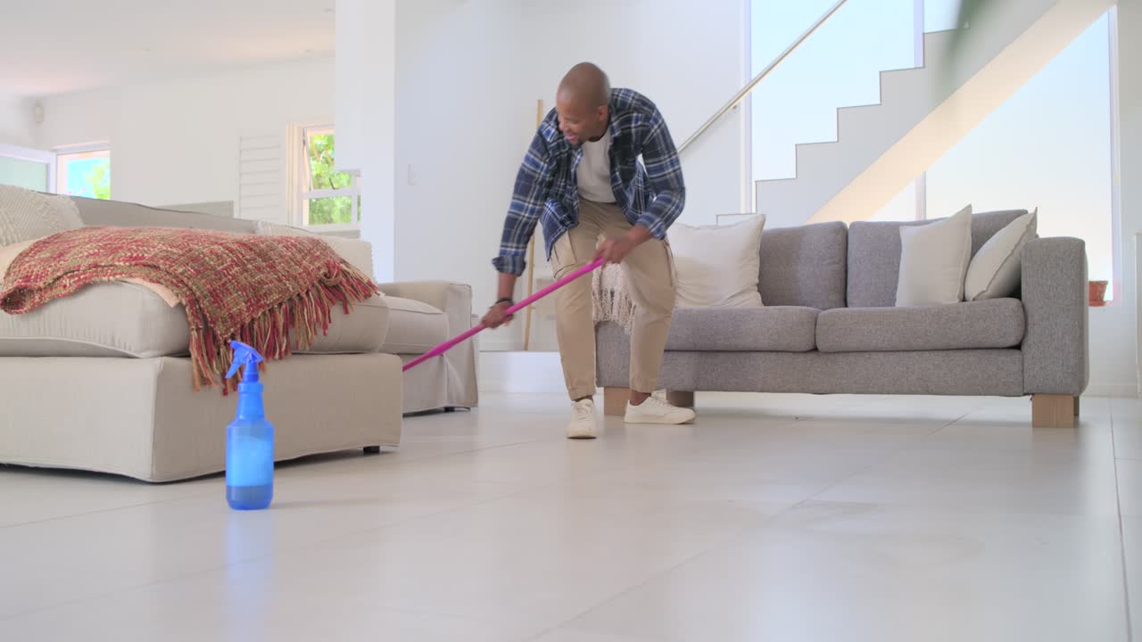 Afroamerikansk mann tar tak i mopp og sprayflaske, mopper stuegulvet for å rengjøre, og sjekker.