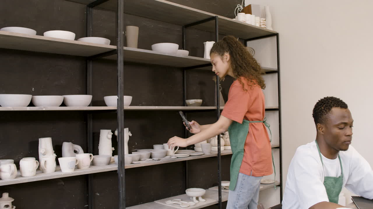 mujer tomando fotos de piezas de cerámica hechas a mano en estantes mientras un colega masculino se sienta en la mesa y trabaja con una computadora portátil en la tienda de cerámica