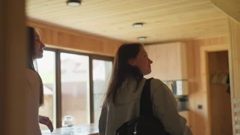 two high school girls step into wooden resort interior, one holding glasses, as they curiously explore space with excitement, soft natural lighting highlighting wooden walls and mounted television