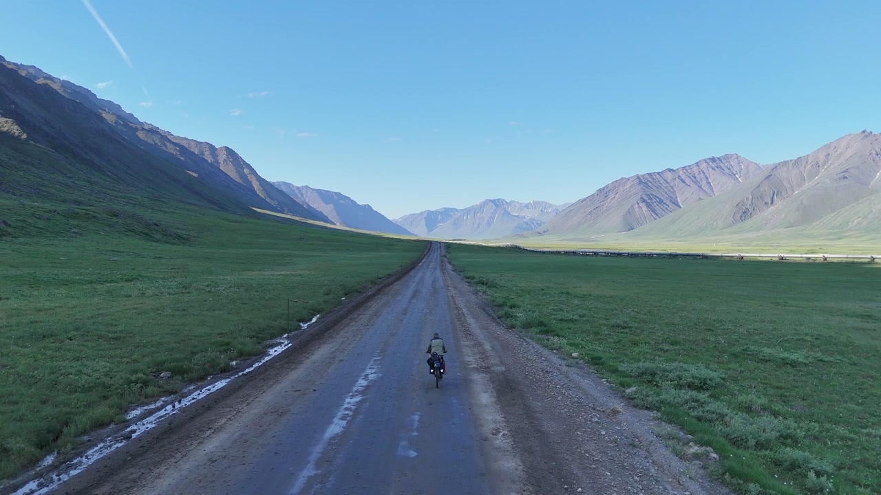 Aerial view of a solitary traveler cycling along a remote dirt road stretching through a vast green mountain valley, representing freedom, adventure, and the spirit of exploration