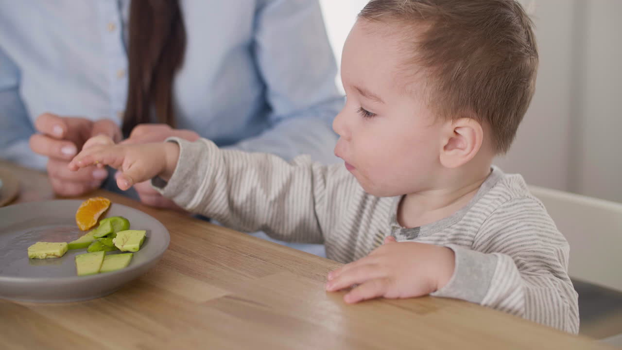 Cute Baby Boy Taking Segment Of Clementine From Plate On Table While His Mom Sitting Next To Him