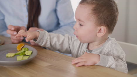 cute baby boy taking segment of clementine from plate on table while his mom sitting next to him