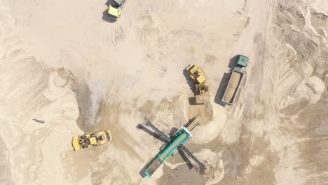 Top-aerial-view-of-bulldozer-loading-sand-into-empty-dump-truck-in-open-air-quarry