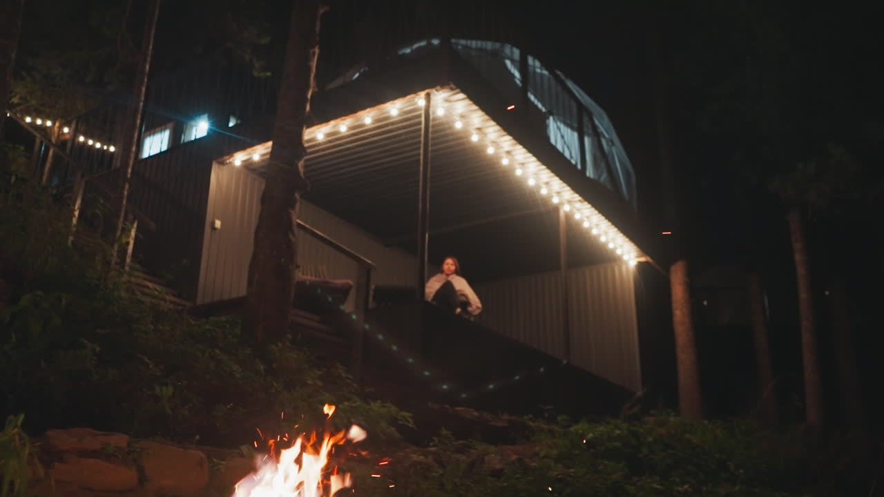 mujer solitaria se sienta en un sillón en la terraza. patio de la casa de glamping con hoguera ardiendo en la noche de verano. relajación en el edificio rodeado de bosque