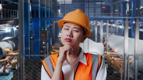 un primer plano de una ingeniera asiática con casco de seguridad de pie en una fábrica de fabricación de turbinas eólicas. pensando en algo y mirando a su alrededor mientras el brazo robótico trabaja