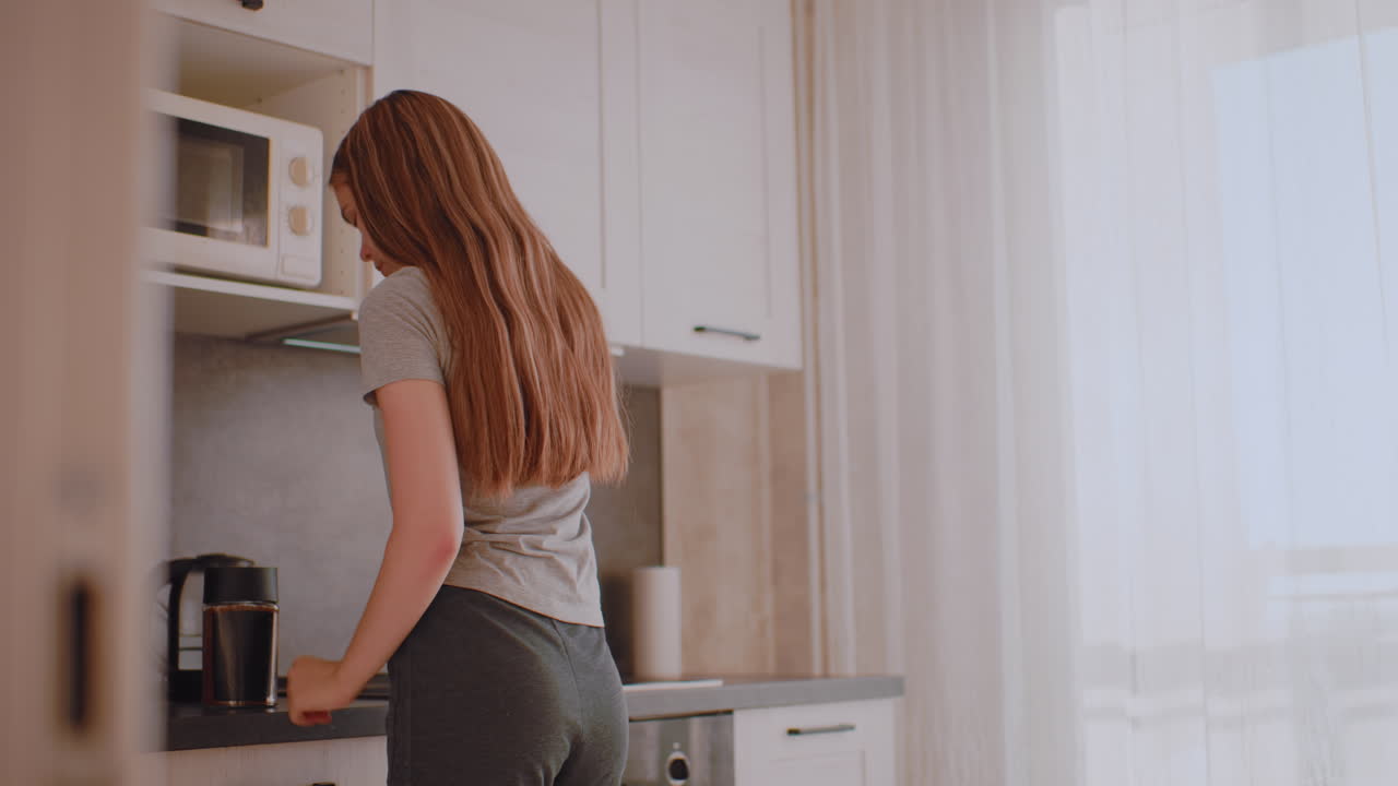 Donna che si prepara per la giornata in cucina: apre il mobiletto per prendere il contenitore del caffè, si prepara a riempire la tazza, apre il mobiletto inferiore per prendere il cucchiaino e inizia a preparare la bevanda mattutina in una calda atmosfera illuminata dalla luce del giorno