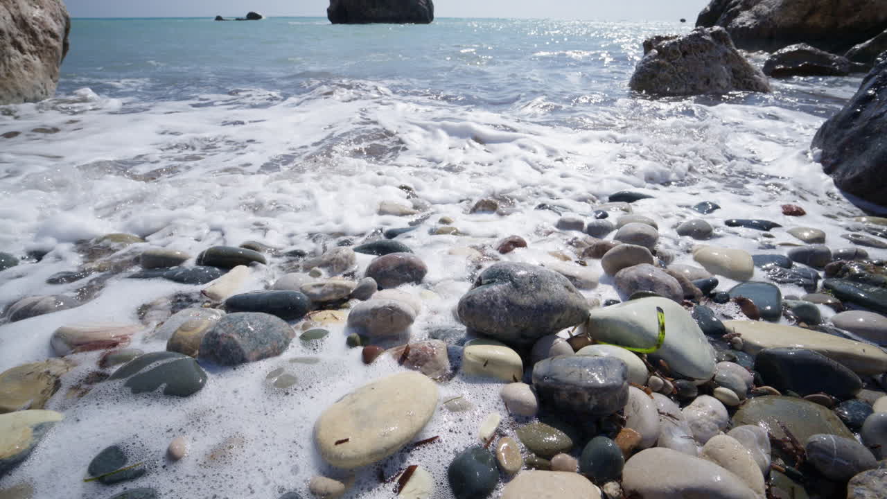 Close up of foamy waves rolling over smooth, multicolored pebbles on the beach near Aphrodite's Rock in Cyprus