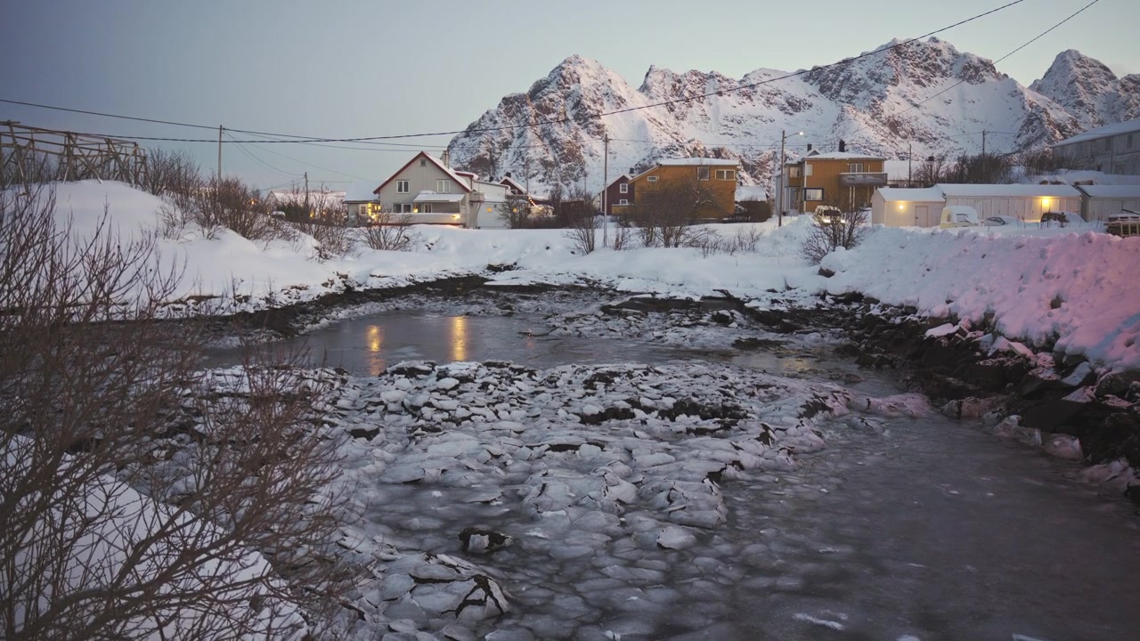 Statisk vidvinkelbilde av en liten frossen innsjø omgitt av tykk snø og rustikke hus i kystbyen Henningsvær om vinteren, Lofoten, Norge.