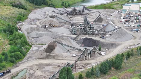 Eine-Große-Kiesproduktionsanlage-Mit-Bulldozern,-Traktoren,-Muldenkippern-Und-Einem-Blick-Auf-Einen-Großen-See-Sowie-Einer-Hochklappbaren-Berglandschaft