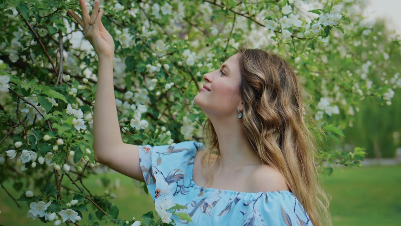 Donna in abito blu tocca rami d'albero in fiore. Sorride dolcemente, con gli occhi chiusi, godendosi la primavera. Una leggera brezza le muove i capelli e i petali. Atmosfera pacifica e romantica.