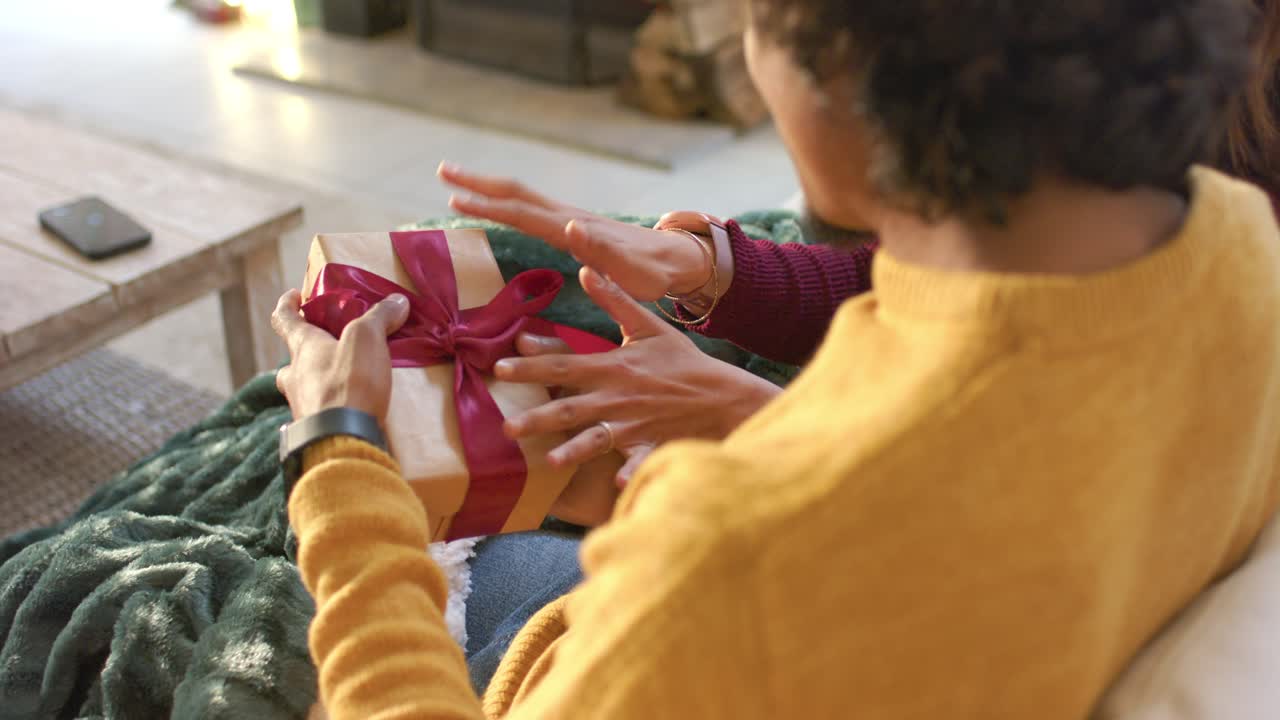 Uomo che offre una scatola regalo dorata mentre una coppia apre un nastro rosso su un divano coperto da una coperta accanto al caminetto.
