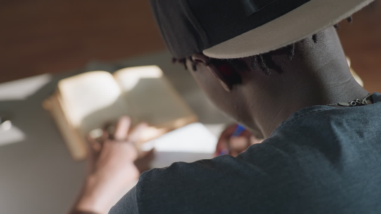 Behind view of man sitting at desk in library, writing notes from open book with pen, focused on study as sunlight illuminates workspace, creating calm and concentrated academic atmosphere