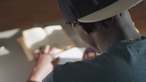 behind view of man sitting at desk in library, writing notes from open book with pen, focused on study as sunlight illuminates workspace, creating calm and concentrated academic atmosphere
