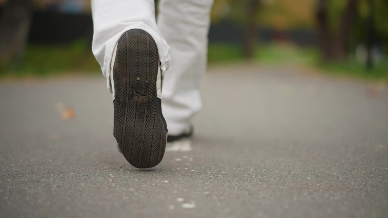 Close up of person legs and feet walking steadily on pavement in outdoor setting. subject wears white pants and dark shoes with camera focused low to capture foot movement against ground