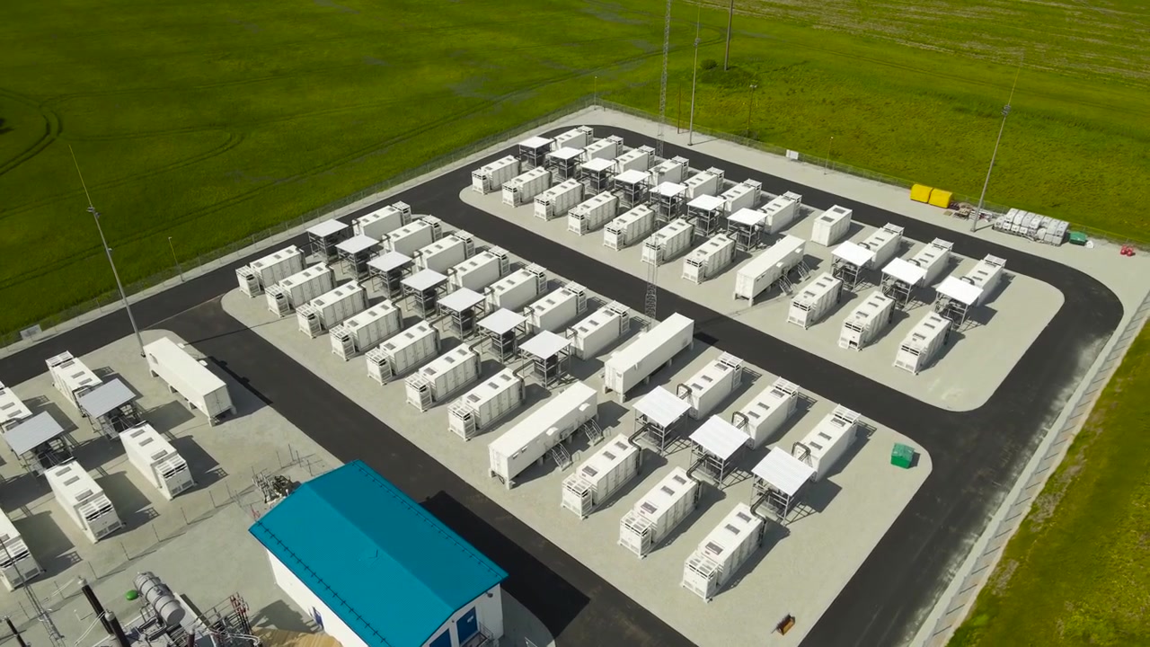 Riprese drone che catturano un moderno impianto di sistema di accumulo di energia a batteria in un campo verde, con file di batterie containerizzate e una sottostazione per la gestione dell'energia rinnovabile.