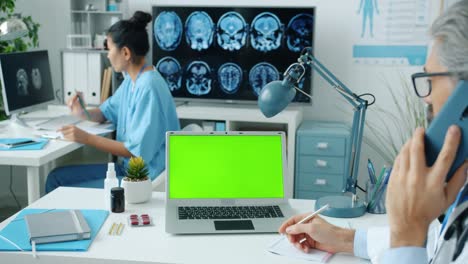 man doctor discussing medicine on mobile phone looking at green chroma key laptop screen in clinic