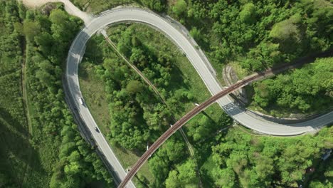 static aerial overhead footage captures multiple cars navigating a winding road intersected by an old railway bridge amidst a forest