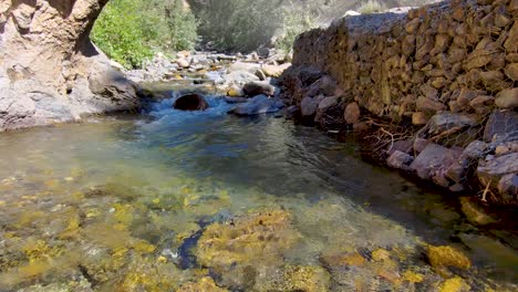 Dolly-forward-just-over-the-surface-of-a-shallow-mountain-creek-flowing-between-a-rugged-cliff-and-a-retaining-wall---aerial-slow-motion