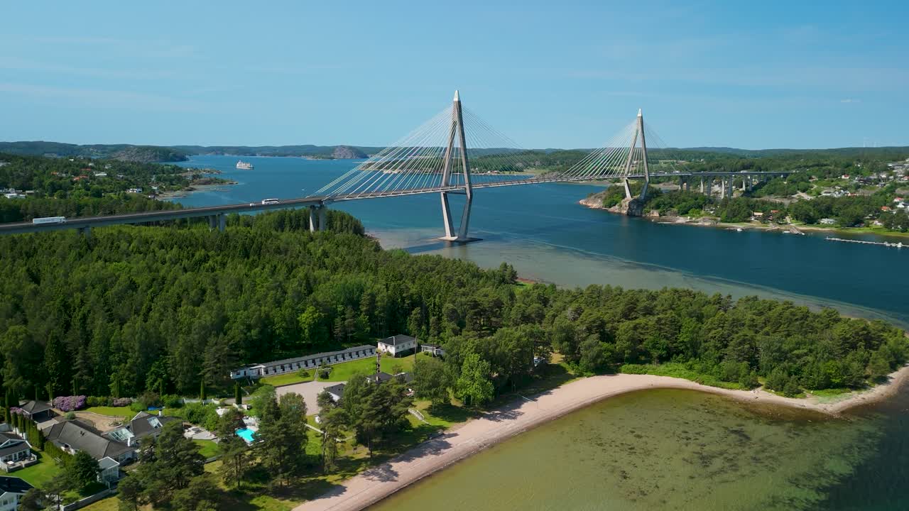Letecké přiblížení dronem k zavěšenému mostu Uddevallabron překlenujícímu fjord Byfjorden s zalesněným poloostrovem, písečnou pláží a panoramatem města Uddevalla v dohledu.