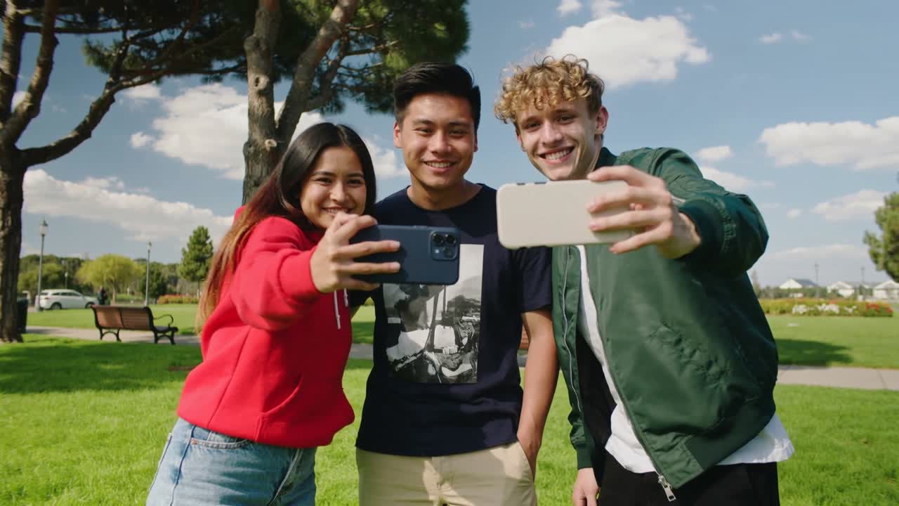 Trio w czerwonych bluzach z kapturem pozuje w parku do selfie, trzymając telefony, jedno z nich wyciąga lewe ramię.