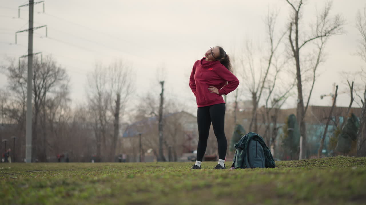 Woman Wearing Hoodie Confidently In Open Space, Young Woman With Backpack Standing Proudly Near Urban Park, Confident Woman Dressed In Red Hoodie Posing Boldly At City Park Entrance