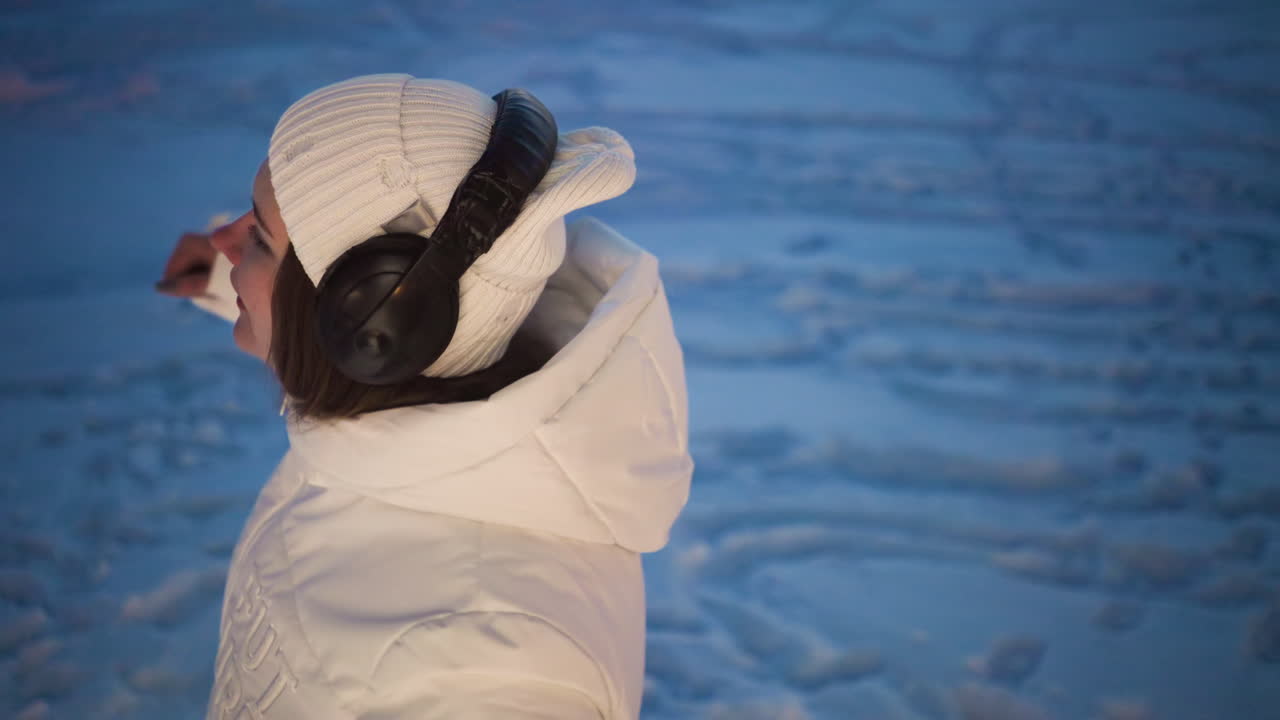 Female artist wearing headphones sways gently in snowy outdoor environment under bare winter trees with rhythmic body movement and soft evening light highlighting flowing coat and crisp winter air