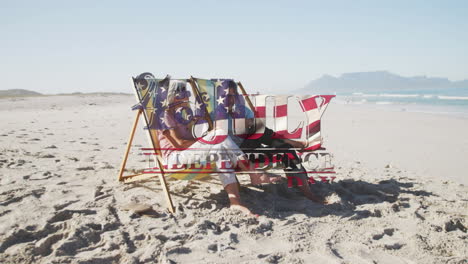 independence day text against senior caucasian couple sitting on deck chairs at beach