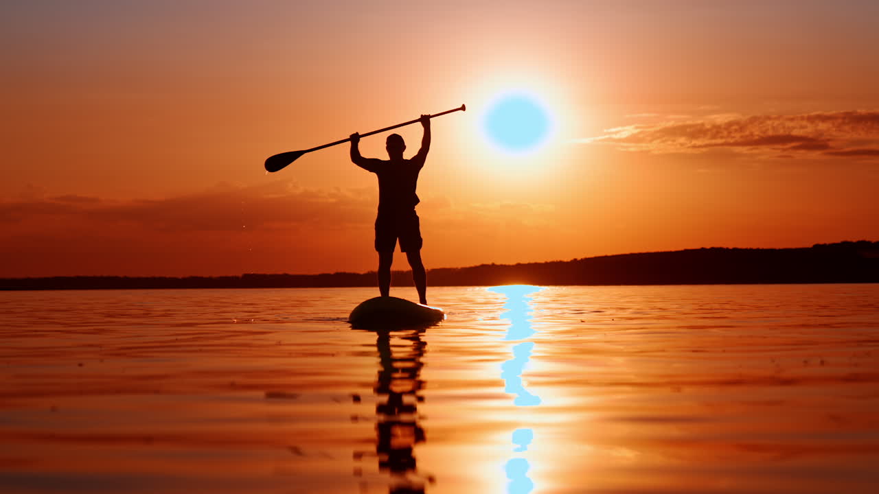 Silhouette di uomo irriconoscibile con cappellino, maglietta e pantaloncini in piedi su una tavola da SUP al tramonto. Lo sportivo ha sollevato il remo sopra la testa.