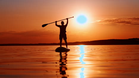 silhouette of unrecognized man in cap, t-shirt and shorts standing on sup board at sunset. sportsman rose the oar over his head.
