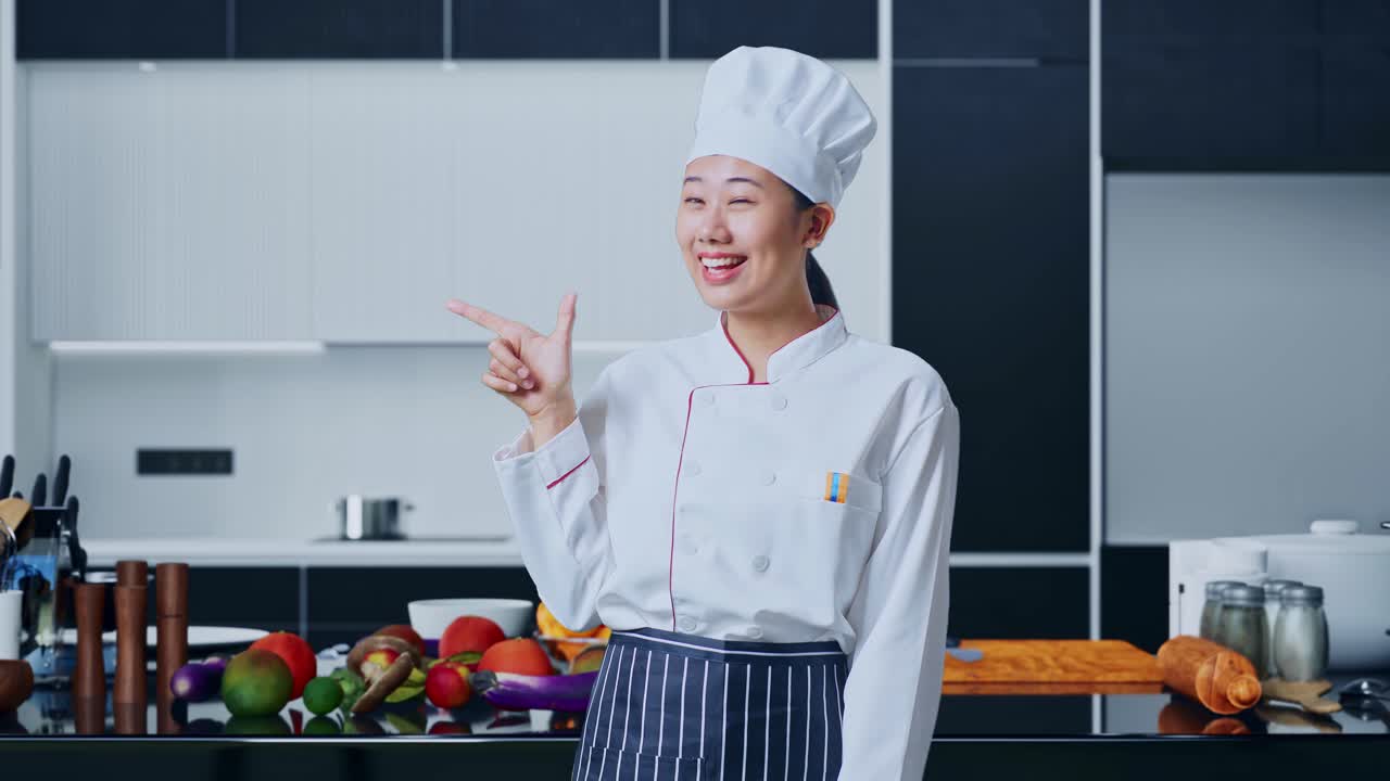 Koki Wanita Asia Tersenyum dan Menunjuk ke Samping di Dapur Rumah