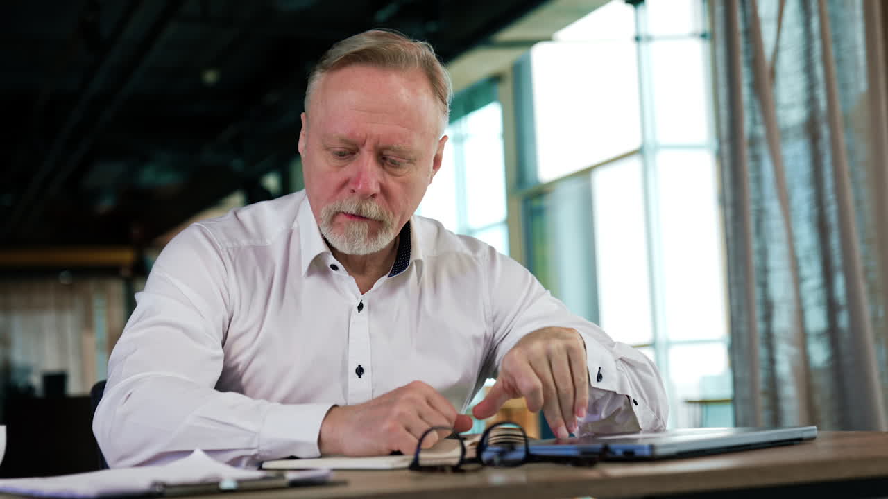 Exhausted businessman sitting at desk closes slowly his laptop. Man puts his glasses to mouth and looks ahead thoughtfully. Low angle view.