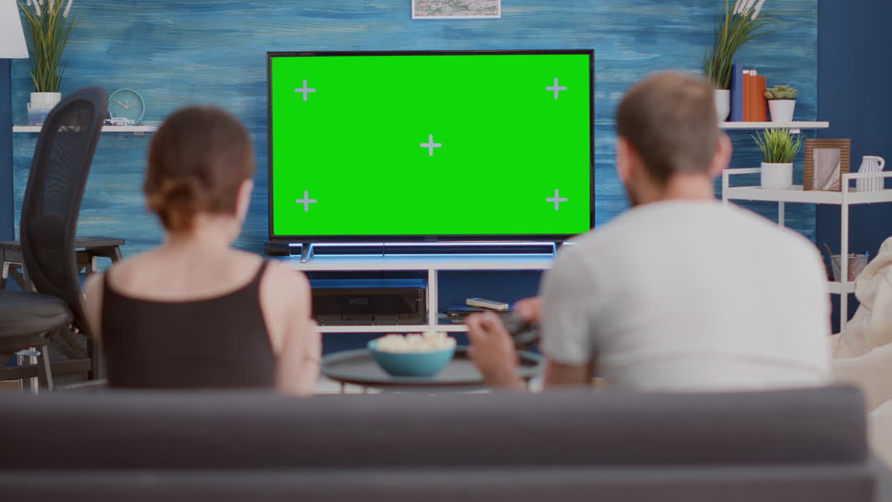 Couple holding controllers playing console online games on green screen tv sitting on couch