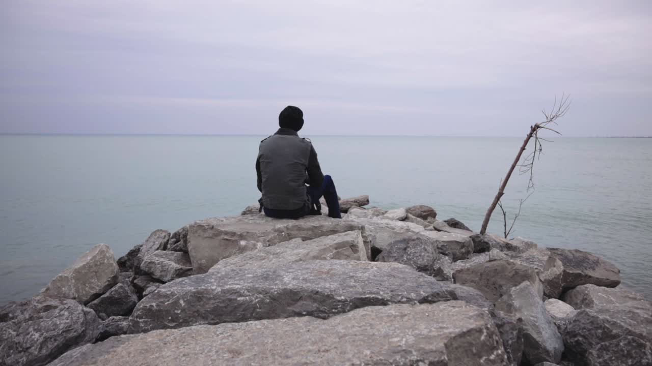 Back View Of A Man Sitting On The Rocky Shore In Canada While Enjoying The Sea - Wide Shot